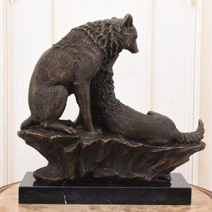 May include: A bronze sculpture of two wolves. One wolf is seated, the other is lying down, both on a rocky base. The sculpture is mounted on a black marble base. The wolves are detailed with textured fur.