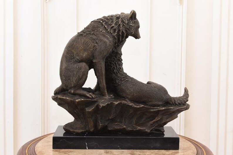 May include: A bronze sculpture of two wolves. One wolf is seated, the other is lying down, both on a rocky base. The sculpture is mounted on a black marble base. The wolves are detailed with textured fur.