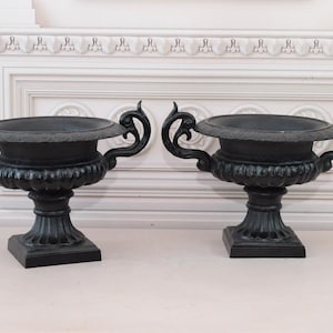 Puede incluir: Dos urnas negras con asas y bases ornamentadas. Las urnas tienen un diseño clásico, con un cuenco ancho y poco profundo y detalles decorativos. Las urnas probablemente están hechas de hierro fundido o un material similar. Adecuadas para uso interior o exterior.