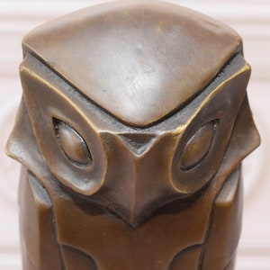 May include: A bronze owl sculpture with a stylized design. The owl's eyes are large and round, and its beak is small and pointed.