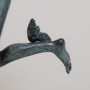 May include: A bronze sculpture fragment depicting a foot and leg with a small wing attached. The sculpture has a verdigris patina, with a detailed rendering of the foot and wing. The background is a plain, light gray.