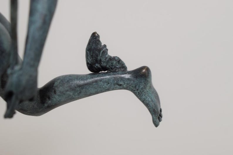 May include: A bronze sculpture fragment depicting a foot and leg with a small wing attached. The sculpture has a verdigris patina, with a detailed rendering of the foot and wing. The background is a plain, light gray.