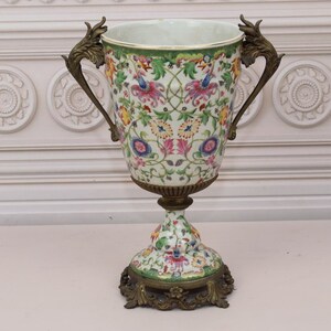 May include: A large white porcelain vase with a floral pattern and gold accents. The vase has two ornate handles and a pedestal base.
