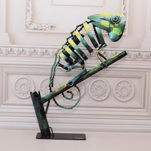 以下が含まれることがあります： 枝に止まっているカメレオンの緑と黄色の金属彫刻。彫刻はリサイクル素材で作られており、ユニークで抽象的なスタイルです。