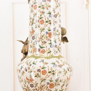 May include: A tall, ornate ceramic vase with a white background and a floral pattern in orange, blue, and purple. The vase has bronze cherub accents and a gold rim and base. The vase is approximately 30 inches tall.