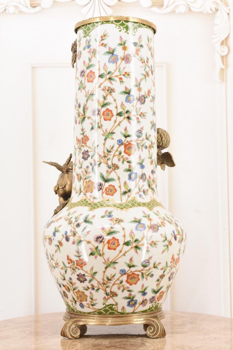 May include: A tall, ornate ceramic vase with a white background and a floral pattern in orange, blue, and purple. The vase has bronze cherub accents and a gold rim and base. The vase is approximately 30 inches tall.