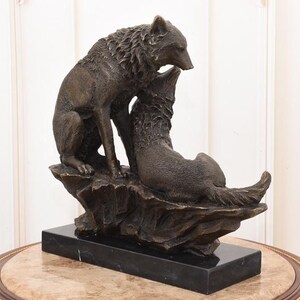 May include: A bronze sculpture of two wolves. One wolf stands over the other, which is lying down. The sculpture is mounted on a black rectangular base. The wolves are detailed with textured fur.