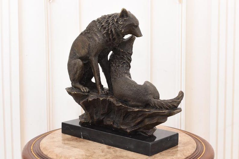 May include: A bronze sculpture of two wolves. One wolf stands over the other, which is lying down. The sculpture is mounted on a black rectangular base. The wolves are detailed with textured fur.