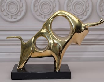 Puede incluir: Una escultura abstracta de un toro de color dorado sobre una base de mármol negro. El toro está estilizado con agujeros en su cuerpo y un cuerno largo y curvo.