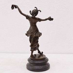 Op de afbeelding: Bronzen beeld van een vrouw in een vloeiende jurk en een hoed, staand op een zwarte basis. De vrouw houdt een stuk stof in haar rechterhand en staat in een dansende pose.