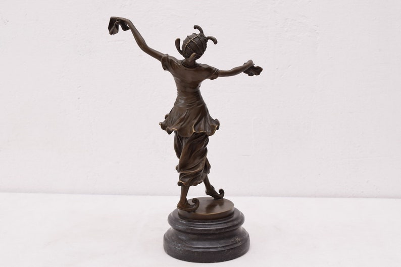 Op de afbeelding: Bronzen beeld van een vrouw in een vloeiende jurk en een hoed, staand op een zwarte basis. De vrouw houdt een stuk stof in haar rechterhand en staat in een dansende pose.