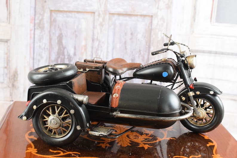 May include: Black and brown vintage-style motorcycle with a sidecar. The motorcycle features a brown leather seat, luggage, and a spare tire. The wheels have a gold finish, and the sidecar has a footrest.