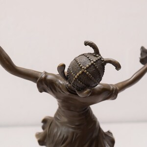 Op de afbeelding: Bronzen beeld van een vrouw die een helm draagt met een decoratief ontwerp. Ze houdt een bloem in elke hand en staat in een danspose.