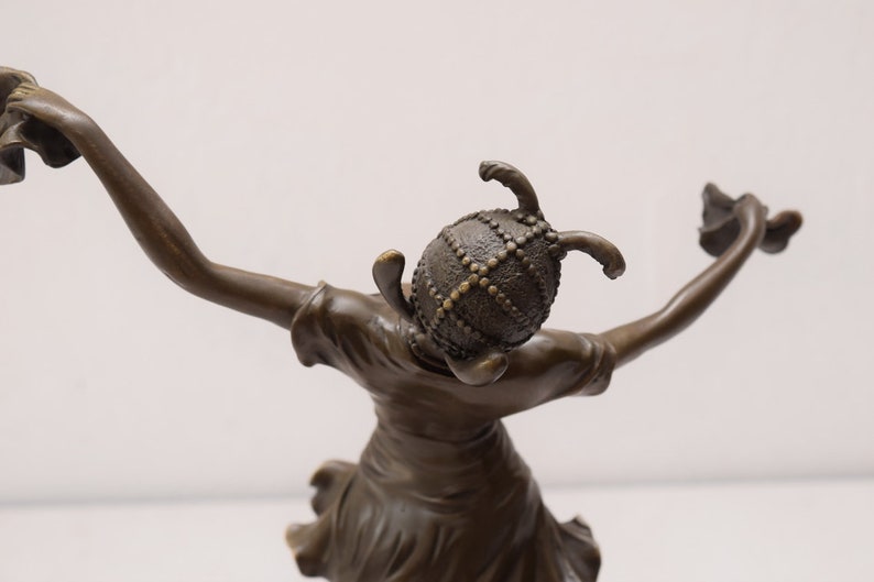 Op de afbeelding: Bronzen beeld van een vrouw die een helm draagt met een decoratief ontwerp. Ze houdt een bloem in elke hand en staat in een danspose.