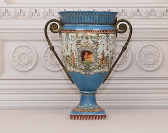 May include: A large, blue and white porcelain vase with gold accents and a decorative floral design. The vase has two brass handles and sits on a white surface.