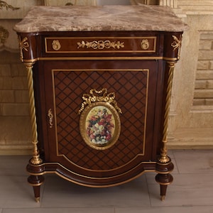Könnte beinhalten: Ein Nachttisch aus dunklem Holz mit Marmorplatte, verziert mit goldfarbenen Metallelementen, einer Schublade und einer Schranktür mit einem zentralen ovalen Blumenmedaillon. Die Tür zeigt ein dunkles Rautenmuster und goldene Zierleisten.