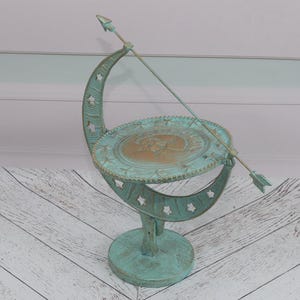 May include: A green metal sundial with an arrow pointing to the time. The sundial is on a white wooden surface.
