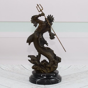Puede incluir: Escultura de bronce de una criatura marina m&iacute;tica, posiblemente Poseid&oacute;n o Neptuno, sosteniendo un tridente. La figura tiene cola de pez y est&aacute; montada sobre una base de m&aacute;rmol negro. La escultura mide aproximadamente 30 cm de altura.