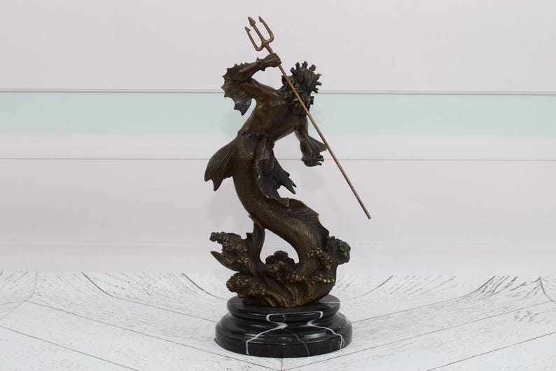 Puede incluir: Escultura de bronce de una criatura marina m&iacute;tica, posiblemente Poseid&oacute;n o Neptuno, sosteniendo un tridente. La figura tiene cola de pez y est&aacute; montada sobre una base de m&aacute;rmol negro. La escultura mide aproximadamente 30 cm de altura.