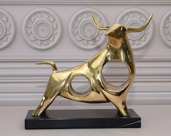 Puede incluir: Una escultura abstracta de toro de color dorado con una base de mármol negro. El toro está estilizado con círculos recortados en su cuerpo.