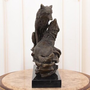 May include: A bronze sculpture of two wolves. One wolf is seated, looking down at the other, which is howling upwards. The sculpture is mounted on a black rectangular base. The wolves are detailed with textured fur.