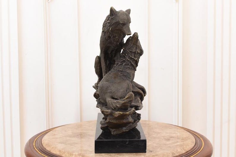 May include: A bronze sculpture of two wolves. One wolf is seated, looking down at the other, which is howling upwards. The sculpture is mounted on a black rectangular base. The wolves are detailed with textured fur.