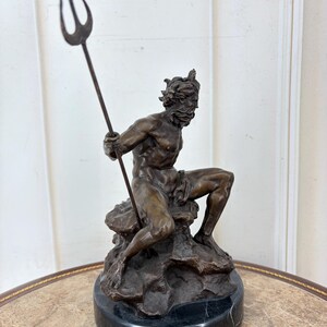 May include: A bronze sculpture of a seated male figure holding a trident. The figure is on a rock formation atop a black marble base. The sculpture has a detailed, aged appearance, with a focus on the muscular physique and facial features.