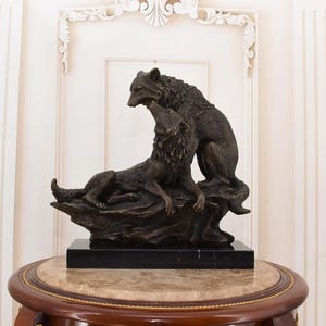May include: A bronze sculpture of two wolves. One wolf is lying down, while the other stands above it. The sculpture is mounted on a black marble base and sits on a round table with a marble top.