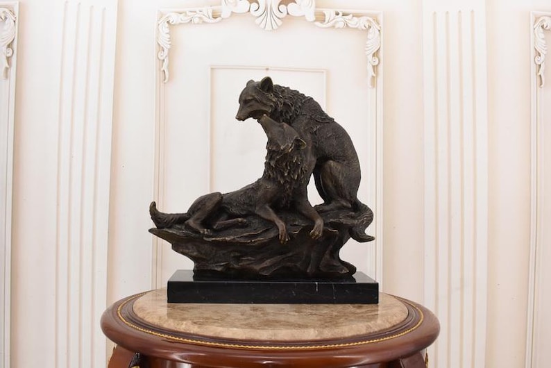 May include: A bronze sculpture of two wolves. One wolf is lying down, while the other stands above it. The sculpture is mounted on a black marble base and sits on a round table with a marble top.