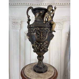 Puede incluir: Una urna de metal en tono bronce oscuro con una figura de querubín sentada en el borde, dos asas ornamentadas y relieves detallados de una lira, cintas y guirnaldas florales. El recipiente se apoya sobre una base con motivos de hojas.