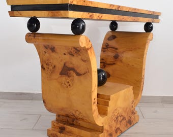 Puede incluir: Una mesa consola con un tablero rectangular de chapa de madera nudosa y una incrustación horizontal negra. La mesa se apoya en dos grandes bases de madera nudosa curvadas en forma de voluta, conectadas por cuatro esferas negras.