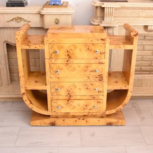 May include: A light brown burl wood chest of drawers featuring four horizontal drawers with small silver-toned knobs. The unit has a curved, U-shaped frame on both sides, creating open shelving sections flanking the central stack of drawers.