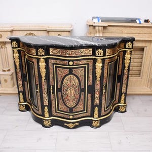 May include: A black and gold cabinet with a marble top. The cabinet has ornate gold detailing and red accents. The cabinet is curved and has glass doors. The cabinet is a piece of furniture.