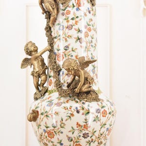 May include: A tall, ornate vase with a white floral pattern and bronze cherub accents. The vase features a gold base and rim, with three cherubs climbing a floral garland. The design includes orange, blue, and purple flowers.