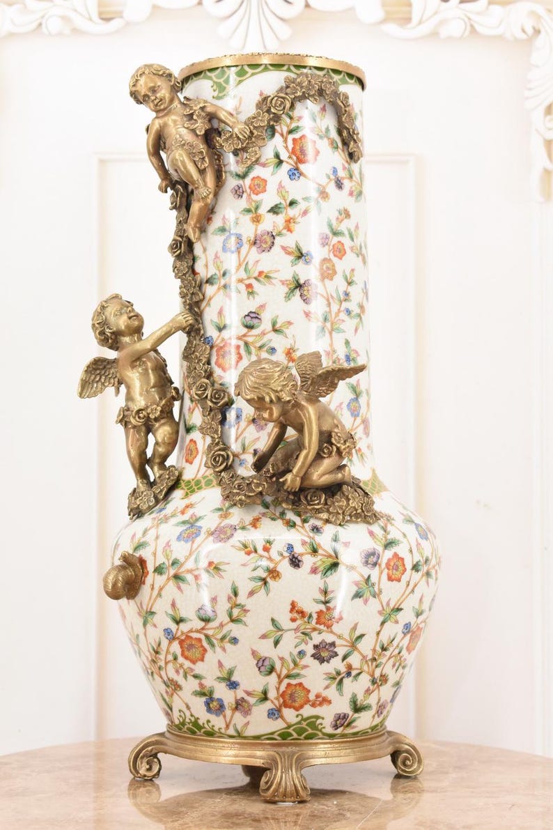 May include: A tall, ornate vase with a white floral pattern and bronze cherub accents. The vase features a gold base and rim, with three cherubs climbing a floral garland. The design includes orange, blue, and purple flowers.