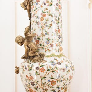 May include: A tall, ornate vase with a white background and a floral pattern in orange, blue, and green. The vase features bronze cherub figures and decorative accents. The base and rim are also bronze.