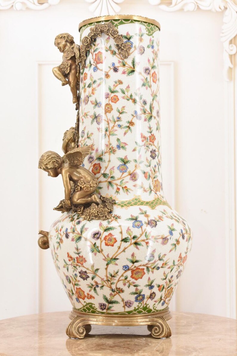 May include: A tall, ornate vase with a white background and a floral pattern in orange, blue, and green. The vase features bronze cherub figures and decorative accents. The base and rim are also bronze.