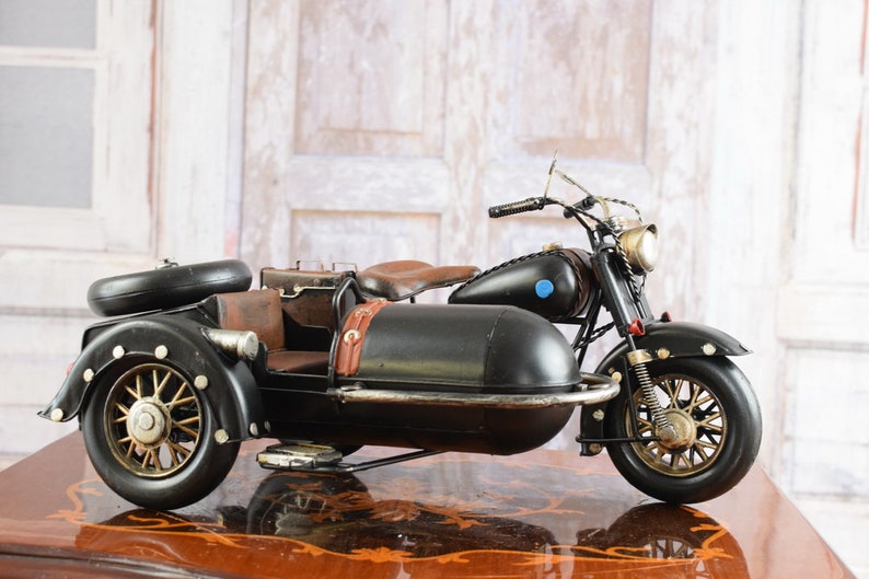 May include: A black and brown vintage motorcycle with a sidecar. The motorcycle has a black body, brown seat, and gold-colored wheels. The sidecar is attached to the right side of the motorcycle. The motorcycle is a decorative item.