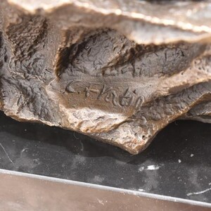 May include: Close-up of a bronze sculpture with a rough, textured surface. The sculpture has a dark brown color with lighter highlights. The artist's signature, "C. Hatin," is visible. The sculpture rests on a black surface.