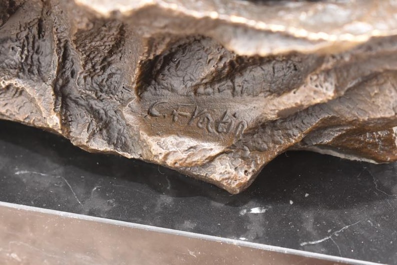 May include: Close-up of a bronze sculpture with a rough, textured surface. The sculpture has a dark brown color with lighter highlights. The artist's signature, "C. Hatin," is visible. The sculpture rests on a black surface.