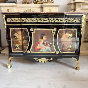 May include: A black commode featuring a pink marble top, gold-toned metal mounts, and two drawers. The central panel displays a painted scene of two figures, while the side panels feature landscape paintings. The piece has four curved legs.