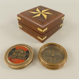 May include: A square wooden box with brass corner accents and a star inlay on the lid, accompanied by a round brass compass and a circular metal tin labeled Eagle Boot Polish Dark Tan with red, black, and gold text and graphics.