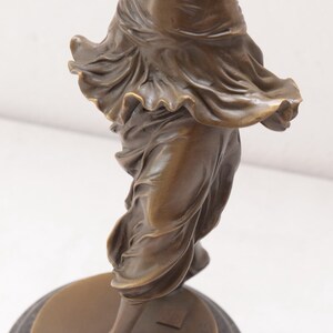 Op de afbeelding: Bronzen beeld van een vrouw in een vloeiende jurk, staand op een ronde basis.