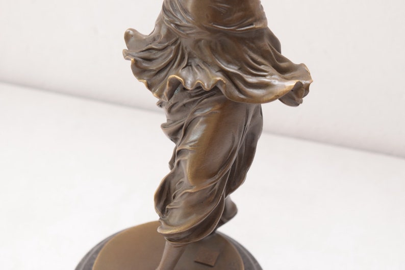 Op de afbeelding: Bronzen beeld van een vrouw in een vloeiende jurk, staand op een ronde basis.