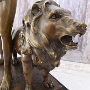 May include: Bronze sculpture of a man standing next to a lion. The man is wearing a loincloth and the lion is roaring with its mouth open. The sculpture is on a black marble base.