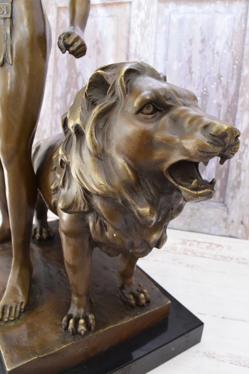 May include: Bronze sculpture of a man standing next to a lion. The man is wearing a loincloth and the lion is roaring with its mouth open. The sculpture is on a black marble base.