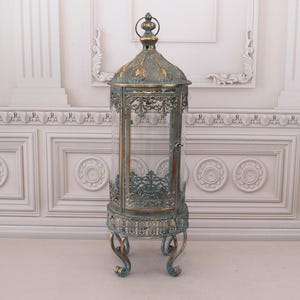 Könnte beinhalten: Eine verzierte, teal-farbene Metalllaterne mit gewölbtem Dach und aufwendigen Filigranarbeiten. Die Laterne hat eine Glaseinfassung und steht auf vier geschwungenen Beinen. Das Design ist vintage-inspiriert und eignet sich für dekorative Zwecke.