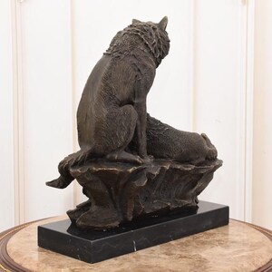May include: A bronze sculpture of two wolves. One wolf is seated, looking upward, while the other rests beside it. The sculpture is mounted on a black rectangular base, and the overall design evokes a sense of wildlife and nature.
