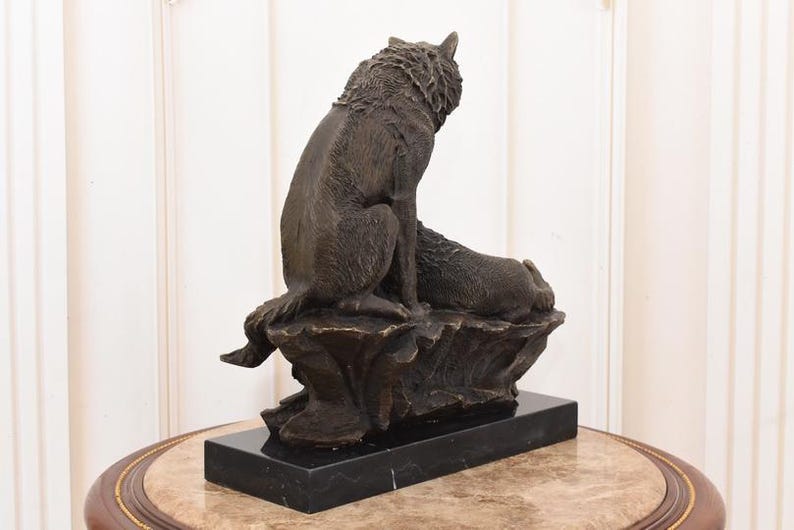May include: A bronze sculpture of two wolves. One wolf is seated, looking upward, while the other rests beside it. The sculpture is mounted on a black rectangular base, and the overall design evokes a sense of wildlife and nature.