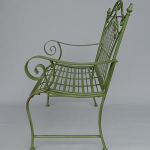 May include: A green metal garden bench with a lattice seat and back. The bench has scrolled armrests and decorative leaf accents on the back. The bench is painted in a distressed, light green color.